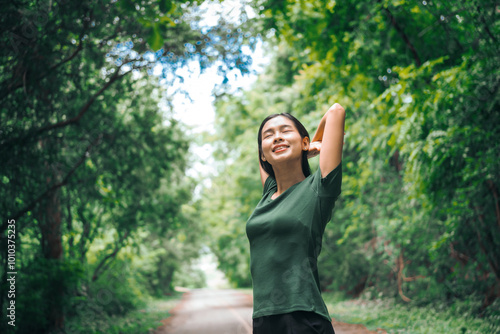 An Asian woman is standing happily in a green forest park, enjoying the fresh air and nature. She takes a deep breath, looks up at the morning sunlight, and feels relaxed and peaceful.