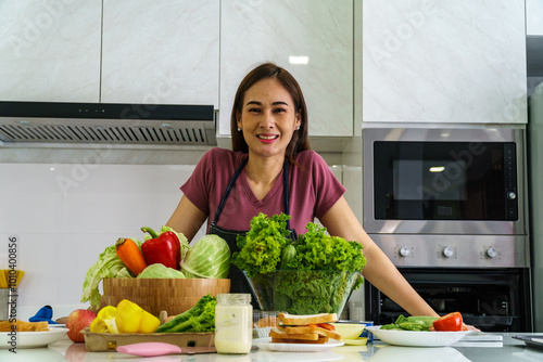 A beautiful Asian woman is standing behind island  of a modern kitchen with fresh vegetables and fruits lined up in front, with a cheerful expression on her face.