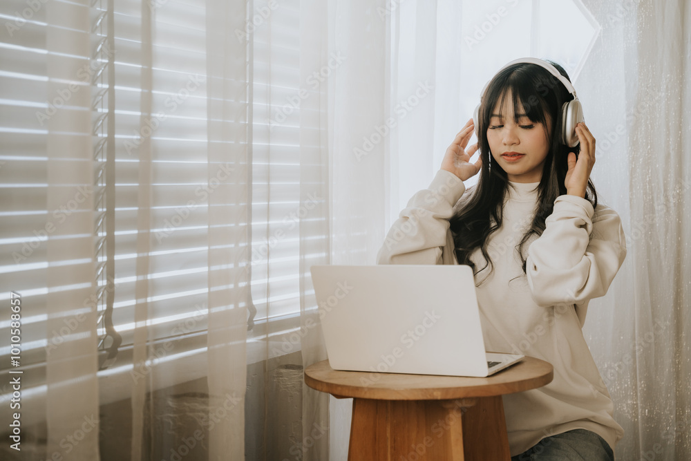 Woman in Cozy Home Office Setting: A young woman wearing headphones sits at a small table, attentively working on her laptop. Natural light streams in from a nearby window. Woman in Cozy Home Office Setting: A young woman wearing headphones sits at a small table, attentively working on her laptop. Natural light streams in from a nearby window.