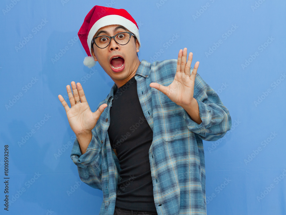 Naklejka premium Portrait of a young Asian man wearing a Santa hat, showing a funny shocked expression while looking forward, isolated against a blue background. Ideal for holiday and seasonal content.