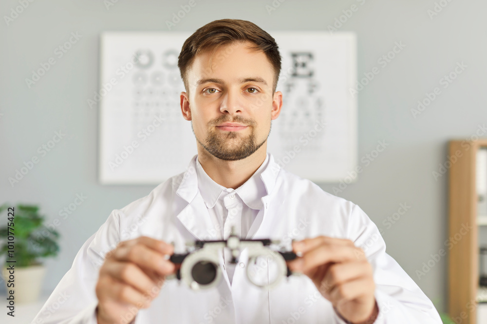 Young male optometrist at work. Portrait of a young man in a white coat holding an optometer, with an eye check chart in the background. Professional optometry, eye health diagnostic concept Young male optometrist at work. Portrait of a young man in a white coat holding an optometer, with an eye check chart in the background. Professional optometry, eye health diagnostic concept