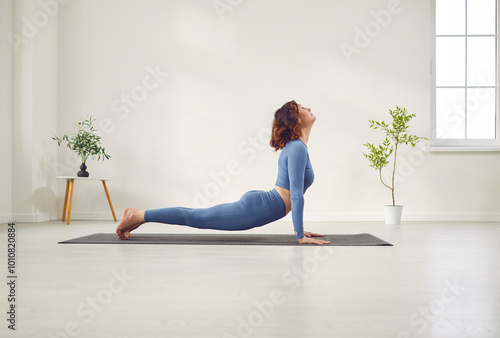 Attractive yogi woman practicing yoga, stretching in Urdhva mukha shvanasana exercise, upward facing dog pose, working out wearing black sportswear in white studio or home room floor background