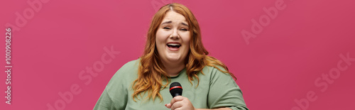 A woman with long red hair sings into a microphone in front of a pink background.