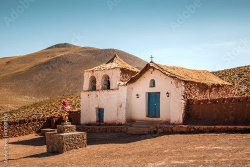 The lovely church of Machuca located in a small village near San Pedro de Atacama in Chile. The church features traditional adobe architecture including thatched roofs and blue doors. photo