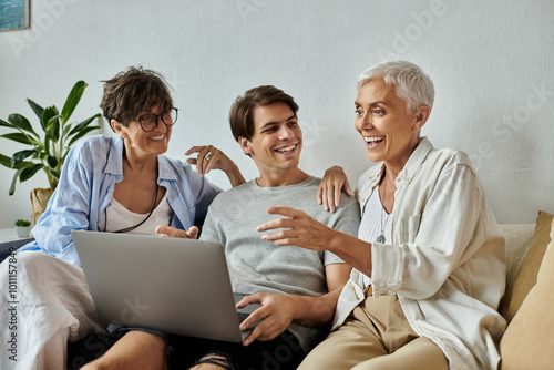 Lesbian parents and their adult son enjoy a joyful moment, sharing laughter and stories around a laptop.