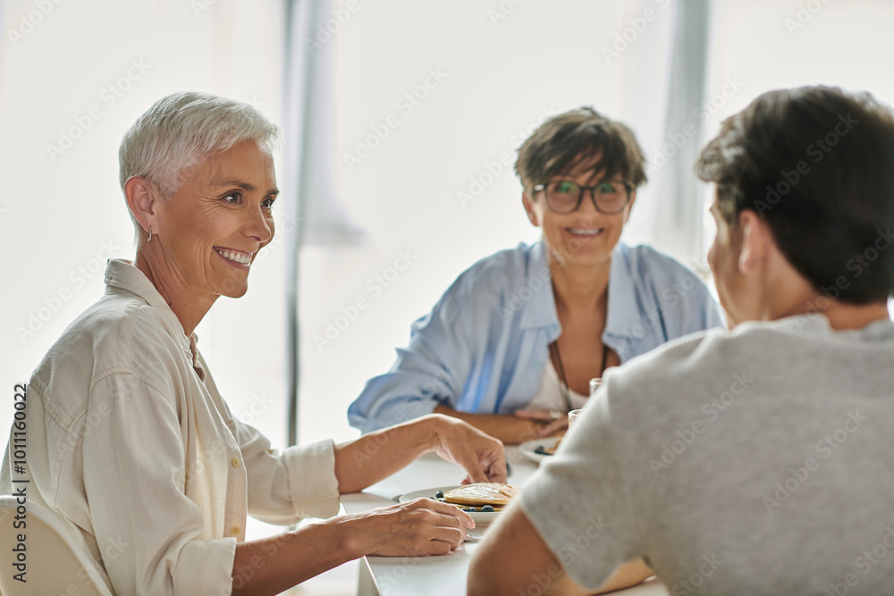 Obraz premium Lesbian parents share a joyful breakfast moment with their adult son at a sunlit kitchen table.