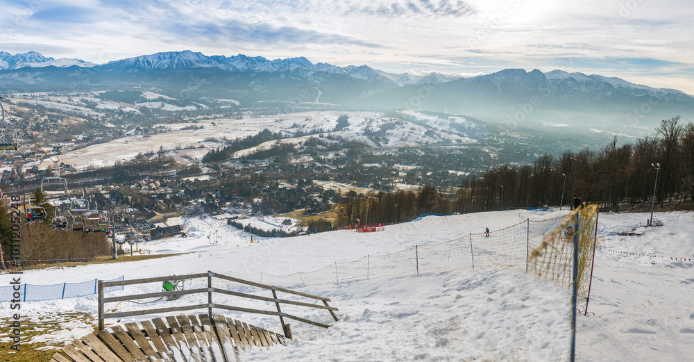 Obraz premium Snowy slopes in Zakopane, Poland, teem with skiers and snowboarders, descending towards a valley lined with scattered buildings