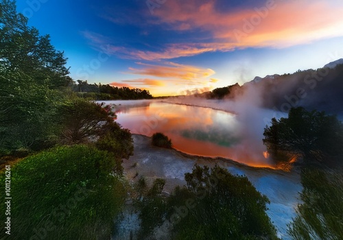 A vibrant New Zealand landscape with a fiery sunrise reflected in a steamy lake.