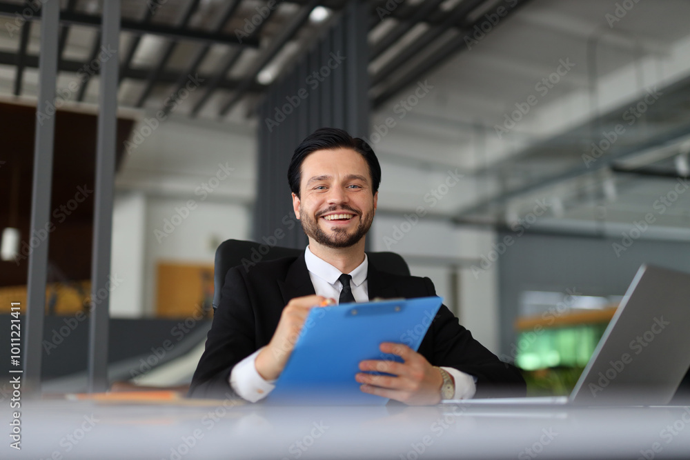 A man in a suit is smiling and holding a blue clip board. He is sitting at a desk with a laptop in front of him A man in a suit is smiling and holding a blue clip board. He is sitting at a desk with a laptop in front of him