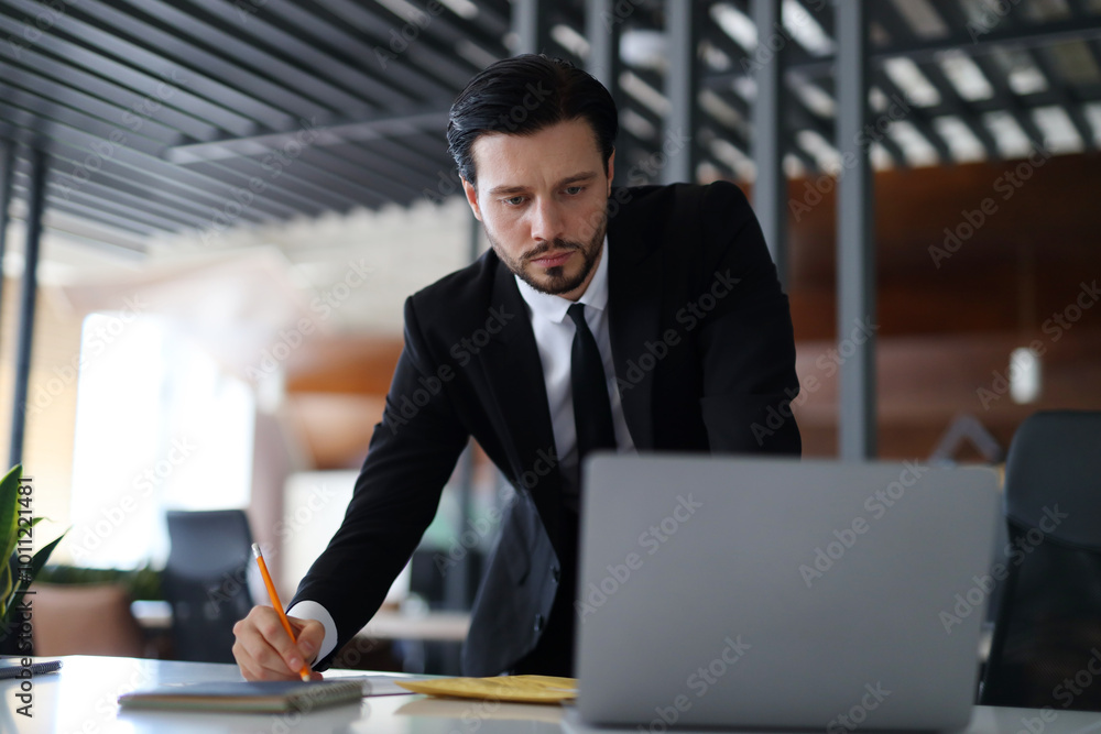 A man in a suit is writing on a piece of paper with a pen. He is looking at a laptop computer A man in a suit is writing on a piece of paper with a pen. He is looking at a laptop computer
