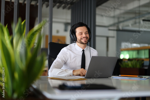 A man wearing a white shirt and a black tie is sitting at a desk with a laptop in front of him. He is smiling and he is enjoying his work photo