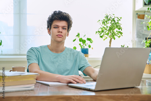 Young male college student sitting at desk with laptop, looking at camera, at home