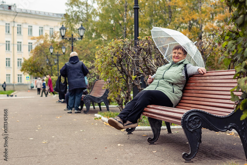 An elderly woman with an umbrella is sitting on a bench.