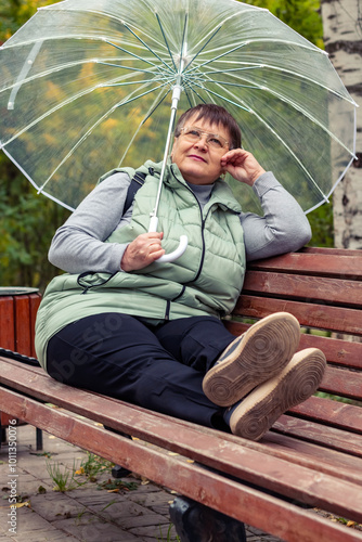 An elderly woman with an umbrella sits on a bench in a city park.