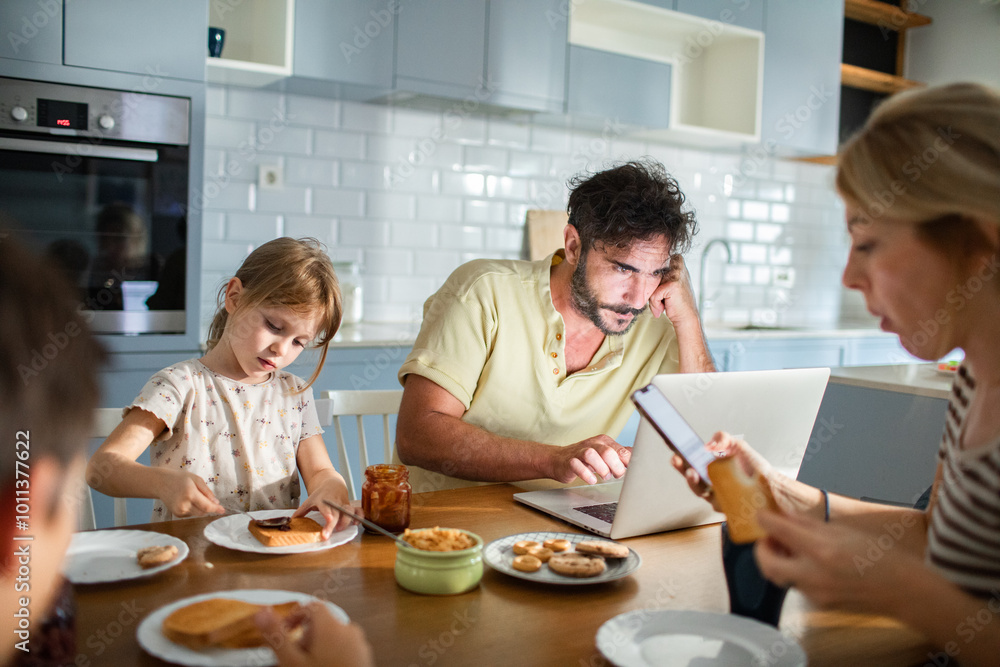 Father working on laptop while family has breakfast in kitchen Father working on laptop while family has breakfast in kitchen