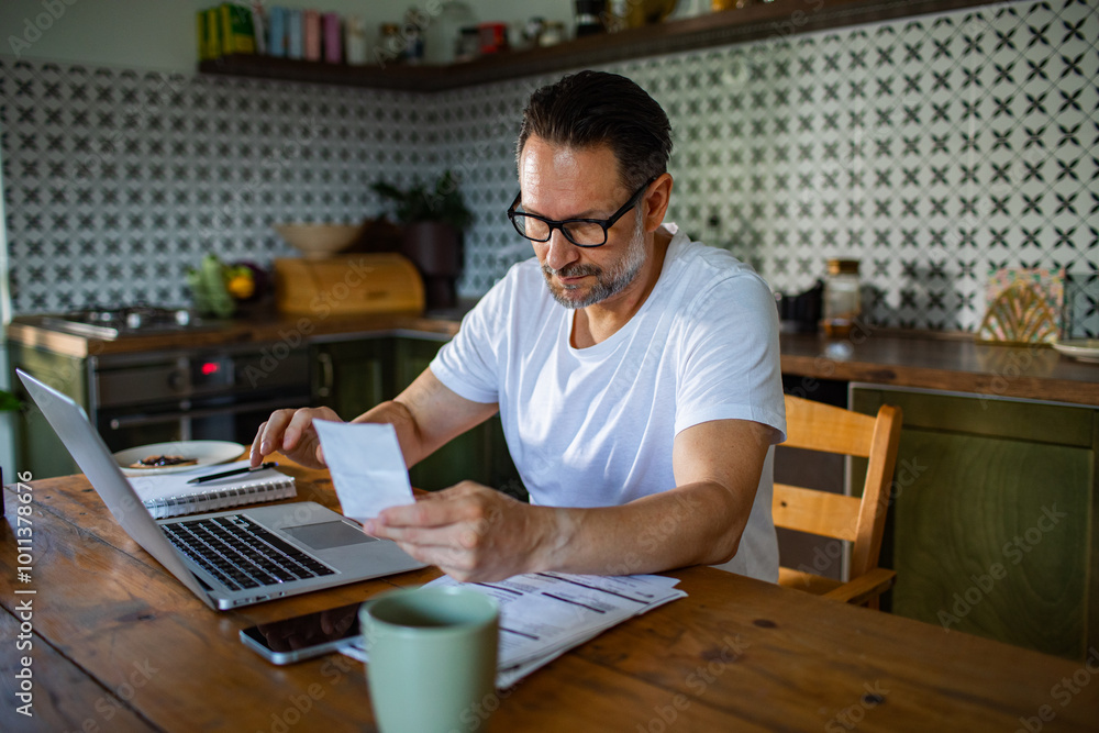 Man reviewing bills and finances on laptop in kitchen Man reviewing bills and finances on laptop in kitchen