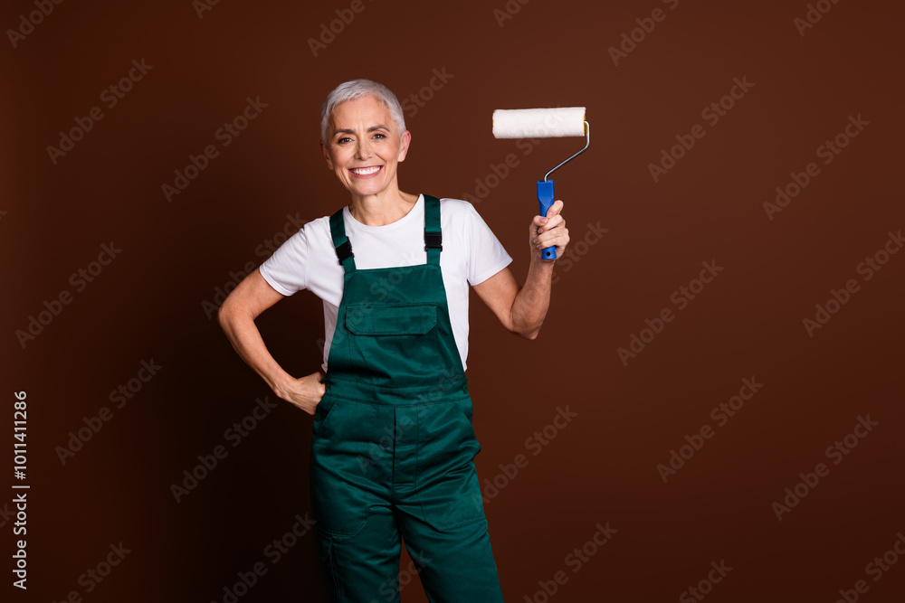 Photo of cheerful pretty lovely senior woman wear green workwear hold brush isolated on brown color background Photo of cheerful pretty lovely senior woman wear green workwear hold brush isolated on brown color background