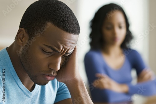 A concerned man with a pensive expression holds his head, while an out-of-focus woman in the background suggests a narrative of worry or conflict.