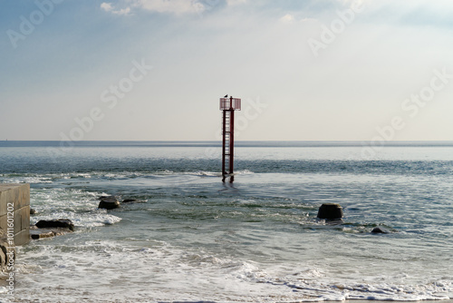 Fin de journée d'été à la barre d'Étel, avec un ciel légèrement voilé et quelques nuages. Une balise indique l'embouchure, et des tétraèdres partiellement immergés ajoutent à cette scène paisible