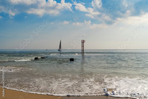 À la barre d'Étel, la fin d'une journée d'été se profile. Un voilier franchit la barre, le ciel est légèrement voilé avec quelques nuages, le sable doré scintille sur la plage, et une balise marque l'