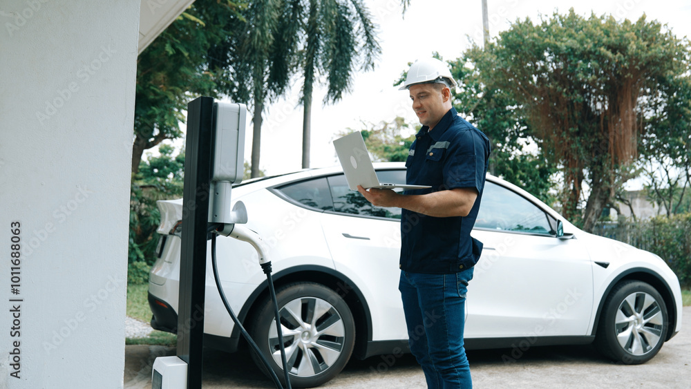 Fototapeta premium Qualified technician working on home EV charging station installation, making troubleshooting and configuration setup on charging system with laptop for EV at home. Synchronos