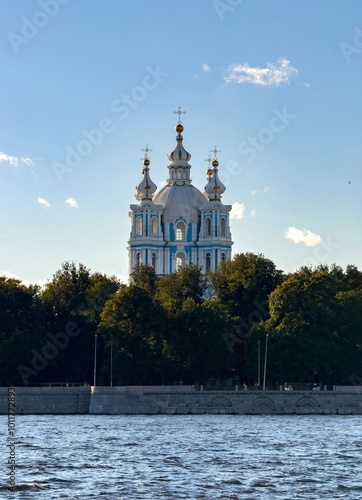A beautiful view of the Smolny Cathedral, standing tall with its golden domes shining in the evening light. The cathedral is framed by lush green trees and a river in the foreground, set against a