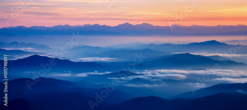 Peaceful panoramic view of rolling mountains covered in mist during sunrise, with soft orange and blue hues filling the sky