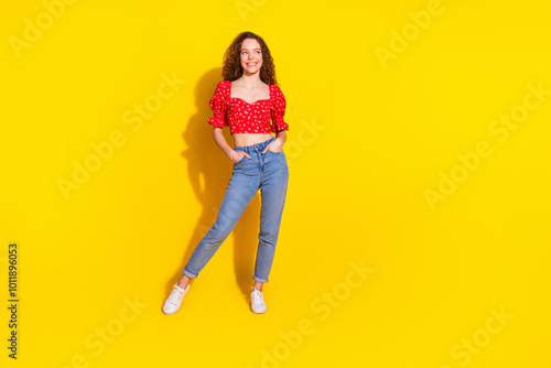 Full size photo of pretty young girl posing hands pockets look empty space wear trendy red outfit isolated on yellow color background