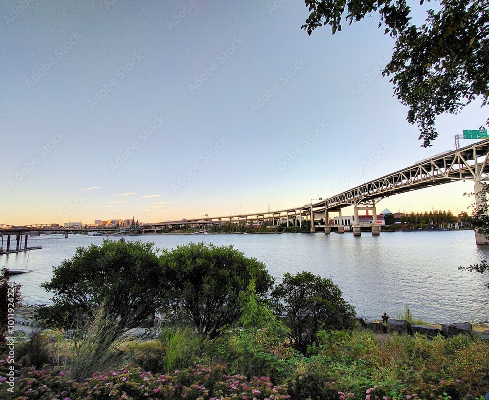 The image captures the Portland road bridge spanning across the calm waters of a river during a serene summer evening in 2023. The sky is clear with soft, warm hues as the sun sets. The image captures the Portland road bridge spanning across the calm waters of a river during a serene summer evening in 2023. The sky is clear with soft, warm hues as the sun sets.