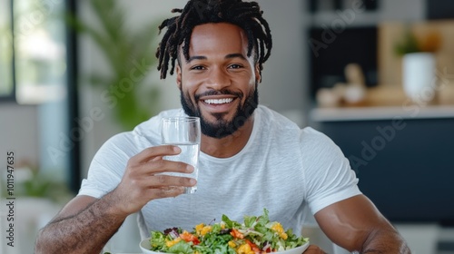 A cheerful man with neatly styled hair and a white T-shirt holds a glass of water, sitting with a vibrant salad placed in front on a sleek dining table. photo