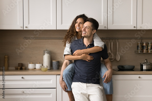 Happy dreamy wife sitting on kitchen counter, embracing husband from behind with love, tenderness. Attractive young couple hugging at home, looking away, talking, planning future