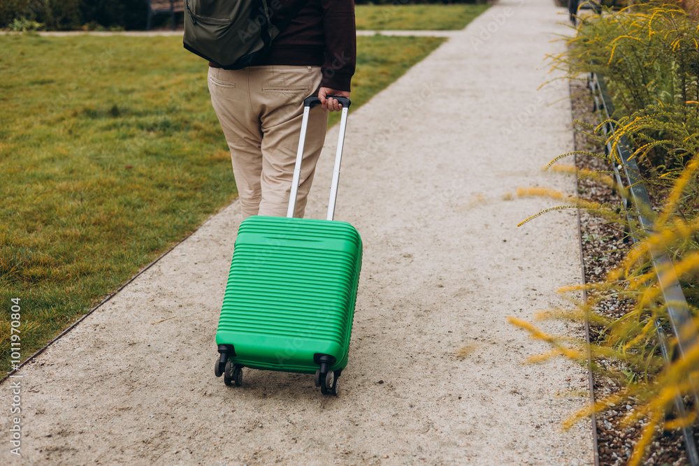Unrecognizable man pulling green suitcase outdoors. Rear view. Copy space. Man walking on the street with travel bag.