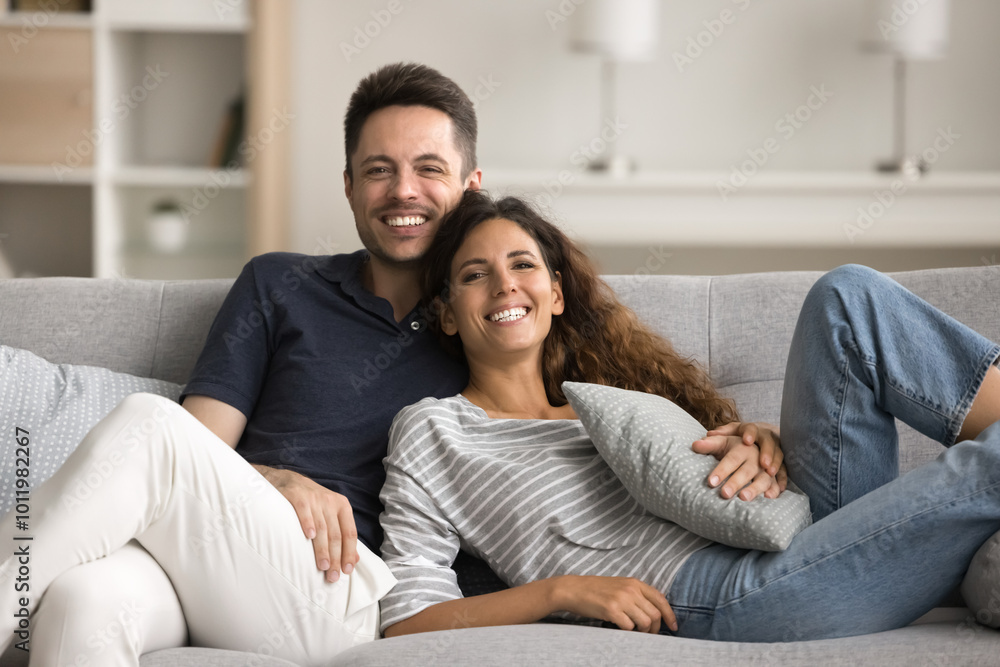 Happy pretty young wife and handsome husband enjoying romantic relationship, leisure on home couch together, hugging with love, affection, looking at camera with toothy smiles Happy pretty young wife and handsome husband enjoying romantic relationship, leisure on home couch together, hugging with love, affection, looking at camera with toothy smiles