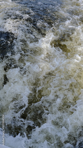 Close-up view of seething river water with white foam. The turbulent current and frothy water create a dynamic texture and energy. A natural scene capturing the power and flow of moving water