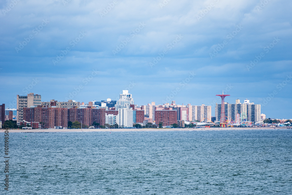 The skyline of Brooklyn emerges from the horizon, seen from a ship sailing through Gravesend Bay. The buildings stand in contrast against the cloudy sky and the steel-colored waters of the bay. The skyline of Brooklyn emerges from the horizon, seen from a ship sailing through Gravesend Bay. The buildings stand in contrast against the cloudy sky and the steel-colored waters of the bay.