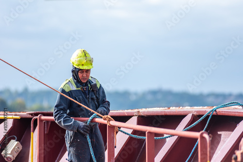 A deck crew member prepares the forward mooring station for docking, focused on securing the ship safely as it approaches the port. The intensity of the task contrasts with the calm surroundings.