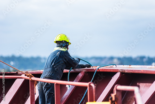A deck crew member prepares the forward mooring station for docking, focused on securing the ship safely as it approaches the port. The intensity of the task contrasts with the calm surroundings.