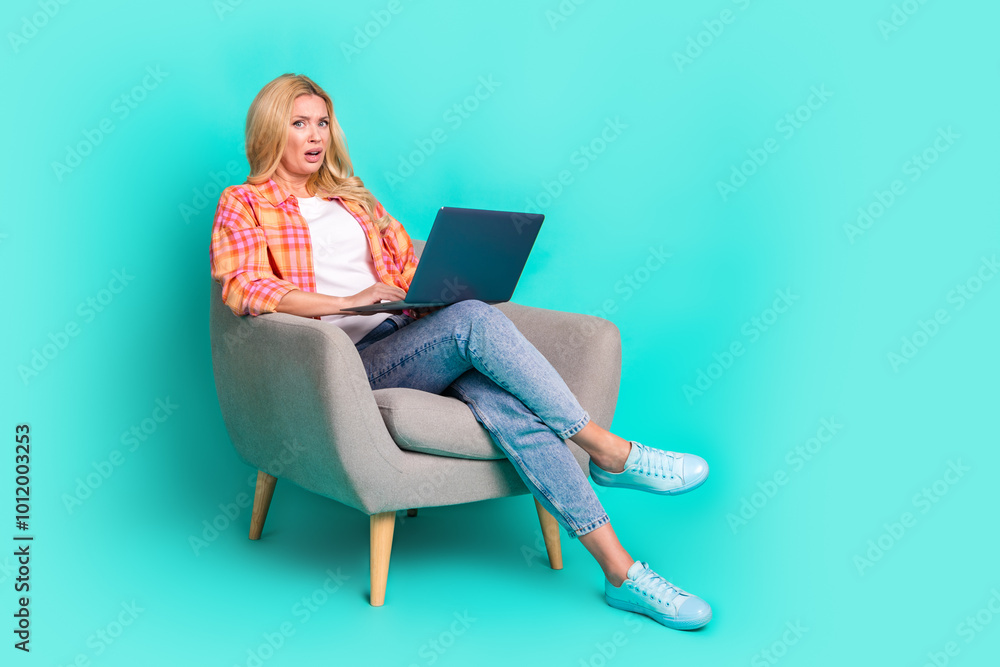 Full body photo of mature woman sit armchair hold netbook shocked scared dressed stylish plaid clothes isolated on cyan color background Full body photo of mature woman sit armchair hold netbook shocked scared dressed stylish plaid clothes isolated on cyan color background