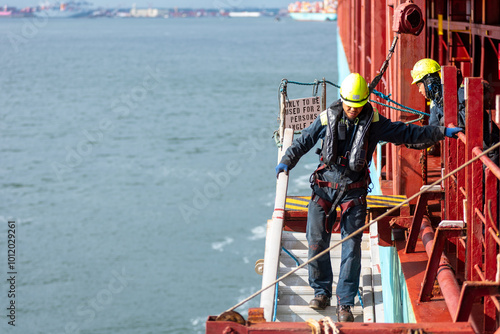 A deckhand prepares the pilot ladder as the ship readies for port entry. Secured in a safety harness, he carefully ensures the setup is ready, focusing on the task in a high-risk environment. 