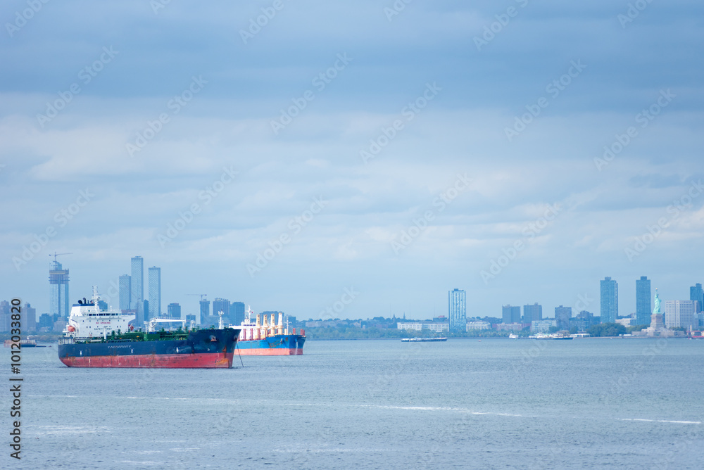 Obraz premium A cargo ship rests anchored in the bay, dominating the foreground. In the distance, the steel-blue skyline of New York City looms under a cloudy sky, blending into the moody, overcast atmosphere.
