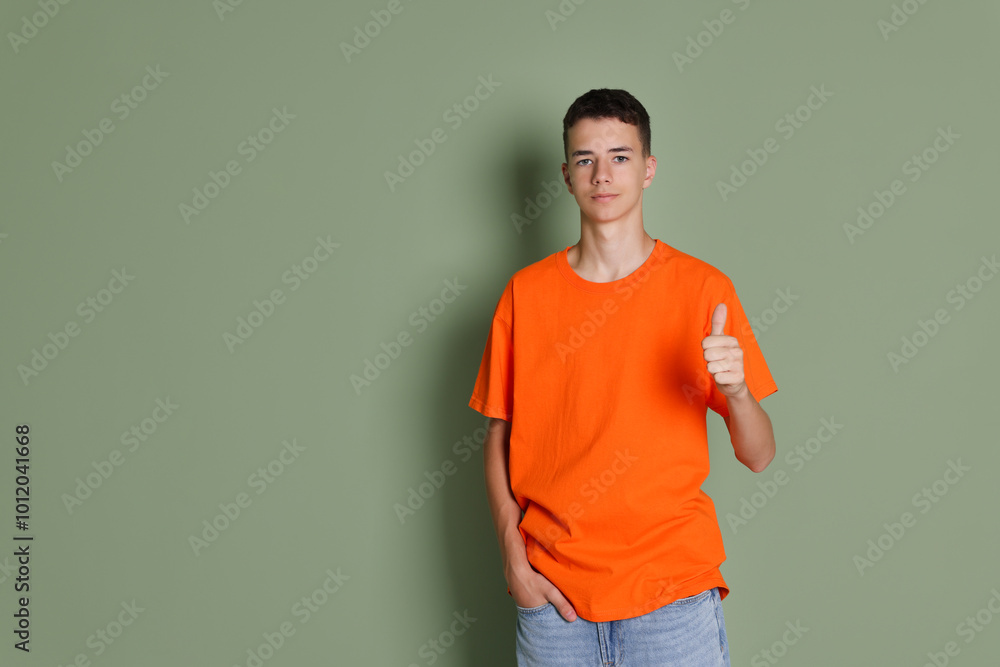 Teenage boy wearing orange t-shirt and showing thumbs up on green background, space for text Teenage boy wearing orange t-shirt and showing thumbs up on green background, space for text