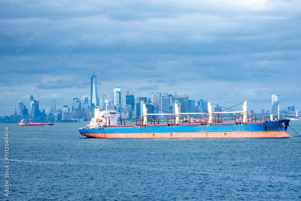 Obraz premium A cargo ship rests anchored in the bay, dominating the foreground. In the distance, the steel-blue skyline of New York City looms under a cloudy sky, blending into the moody, overcast atmosphere.