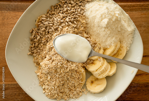 Close up of a deep plate with muesli ingredients, as oatmeal, sunflower seeds, banana slices, protein powder and a spoonful of yogurt. Table stands on a wooden snackboard. photo