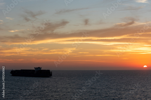 The calm silhouette of a massive container ship gliding across the smooth sea, enveloped by the golden hues of the sky as the sun bids farewell to the day.