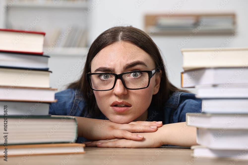 Worried student before exam at table among stacks of books indoors Worried student before exam at table among stacks of books indoors