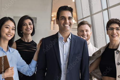 Positive successful male Indian boss posing for portrait with diverse employees, looking away with toothy smile, enjoying leadership, teamwork, feeling team support, unity