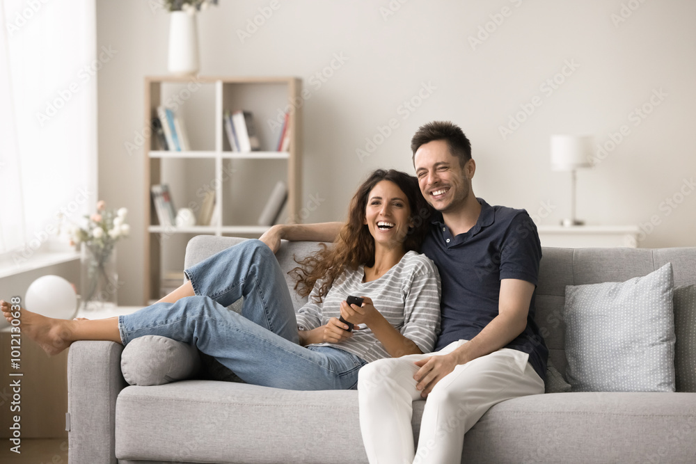 Joyful excited pretty couple spending leisure time at TV, using remote control, resting on comfortable home sofa, watching movie, sitcom show, comedy, online channel, hugging, laughing Joyful excited pretty couple spending leisure time at TV, using remote control, resting on comfortable home sofa, watching movie, sitcom show, comedy, online channel, hugging, laughing