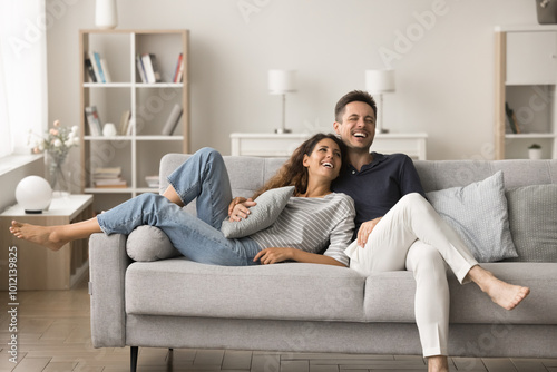 Positive happy young husband and wife relaxing on new comfortable pale grey sofa, hugging, talking, laughing, having fun, spending home funny leisure time together photo