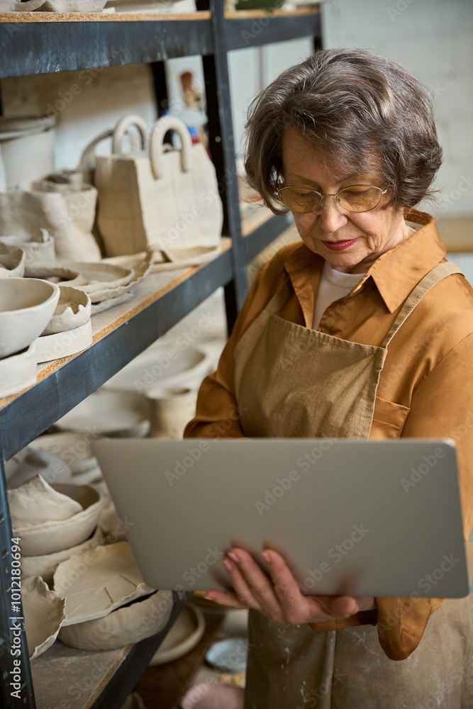 Fototapeta premium Lady with laptop stands by shelf with unfired clay products