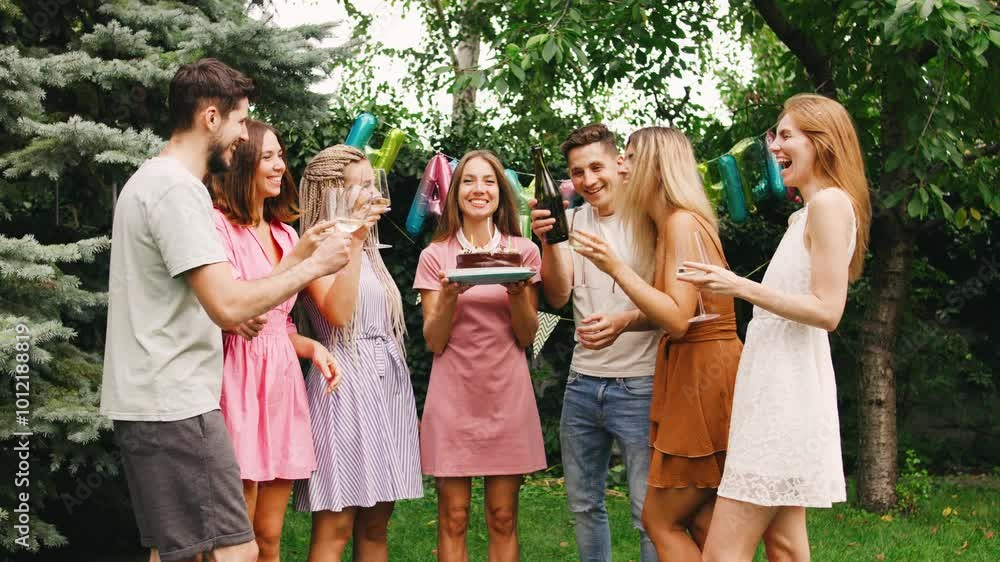 Woman with birthday cake with lit candles stands surrounded by her friends in front of festive decorations Woman with birthday cake with lit candles stands surrounded by her friends in front of festive decorations